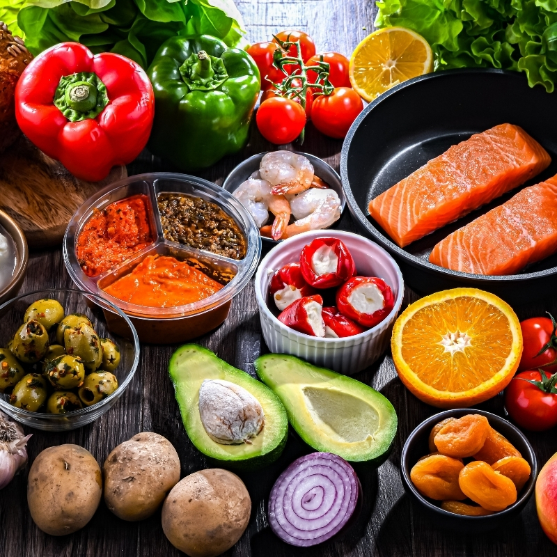 A variety of fresh, colorful foods including salmon, shrimp, avocado, vegetables, fruits, and healthy fats arranged on a table, representing a balanced and nutritious Mediterranean diet.